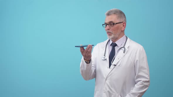 Doctor Man Communicates at Phone and Correcting Glasses Stands in Studio with a Blue Background