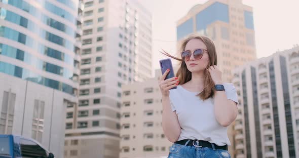 Young Beautiful Woman Smiling with Smartphone in Hand Against Summer Skyscrapers