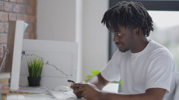 Happy Smiling Man Texting on Smartphone Looking at Computer Monitor Screen Sitting at Table Indoors