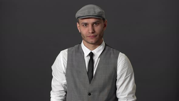 Portrait of Pleased Mature Guy in Vest and Flat Kepi Smiling and Showing Thumb Up Over Gray