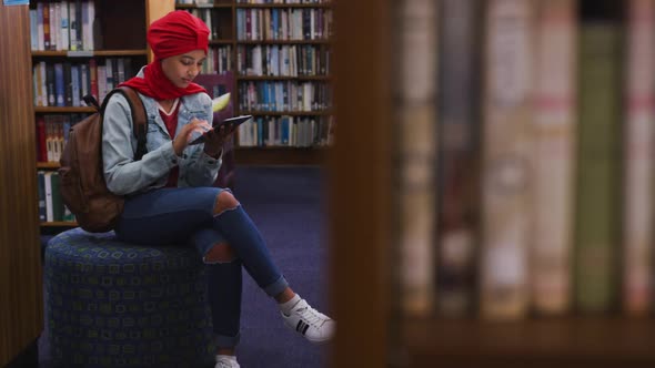 An Asian female student wearing a red hijab studying in a library and using tablet