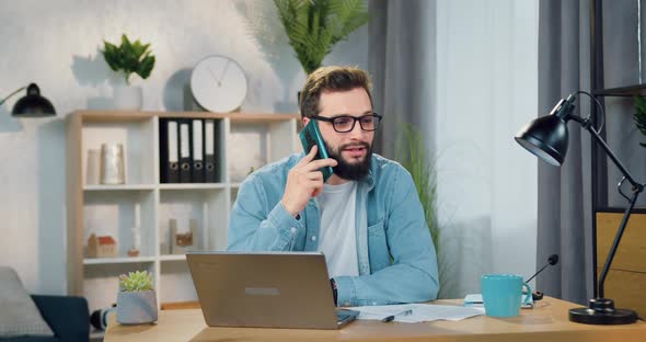Man in Glasses Talking on Smartphone while Working on Laptop in Beautifully Decorated Room