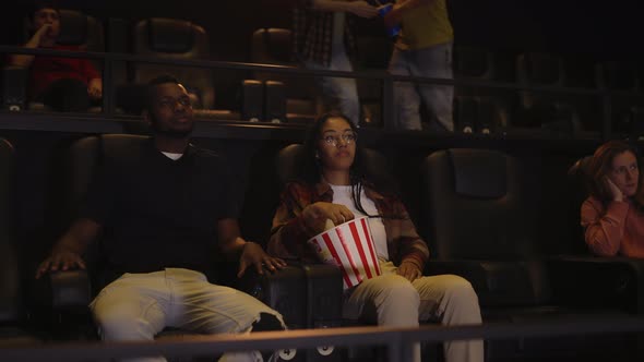 African American Young Couple Sitting in Movie Theatre Watching Film and Eating Popcorn