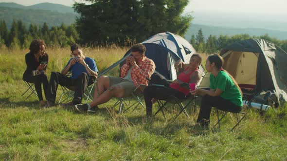 Chatting Diverse Multiracial Backpackers Relaxing and Drinking Hot Drinks in Morning at Campsite