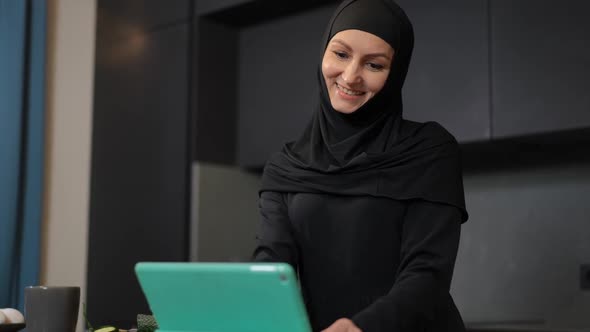 Beautiful Middle Eastern Woman Messaging Online on Tablet Smiling Standing in Kitchen at Home