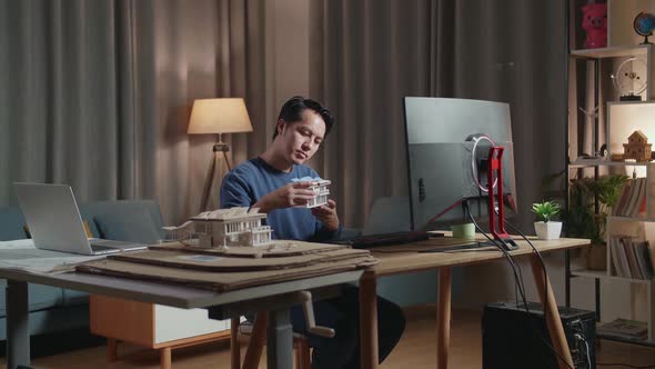 Asian Male Engineer Holding And Looking At House Model Before Comparing To Design in Desktop At Home