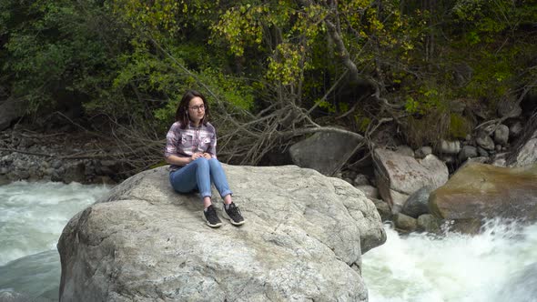 A Young Woman with Glasses Sits on a Stone with Headphones in the Mountains Near the River. Travel