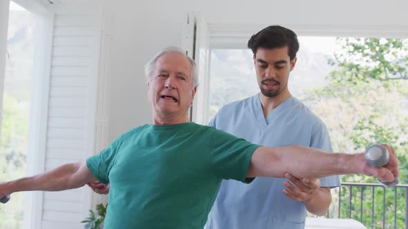 Young male physiotherapist helping retired senior man exercising with dumbbells at nursing home