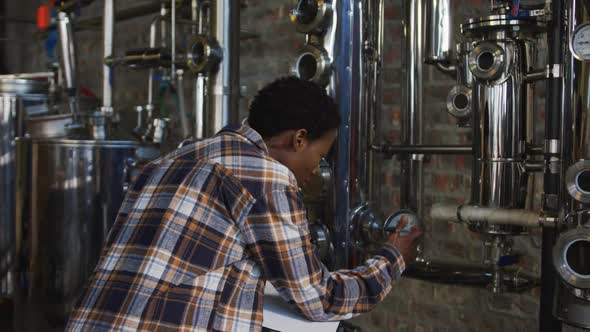 African american woman working at gin distillery checking equipment and writing on clipboard