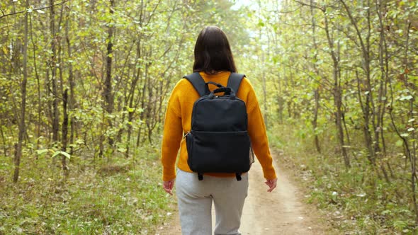 Woman with Backpack in Orange Sweater Walks Across Forest