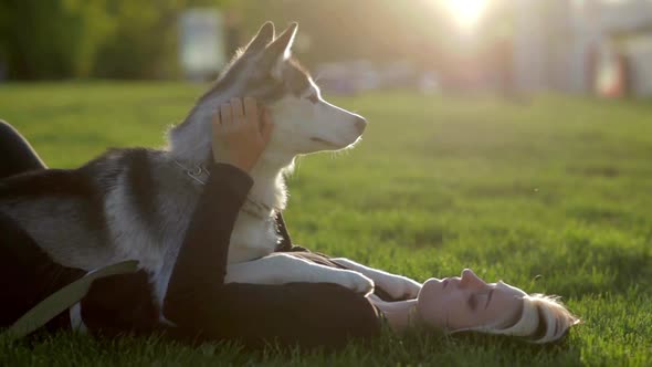 Beautiful Young Woman Playing with Funny Husky Dog Outdoors in Park at Sunset or Sunrise on Green