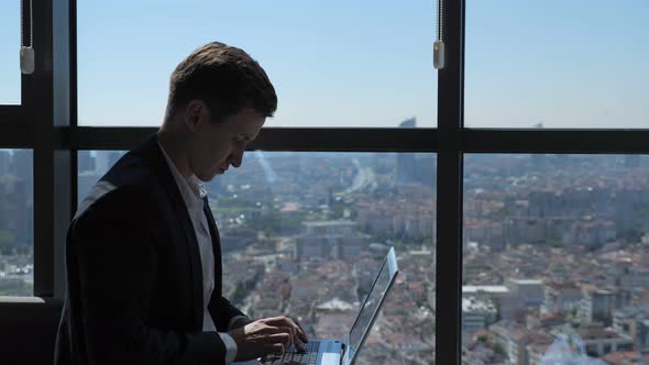 Ofiice Worker in Suit Is Typing on Laptop Sitting Near the Window with Panoramic City View.