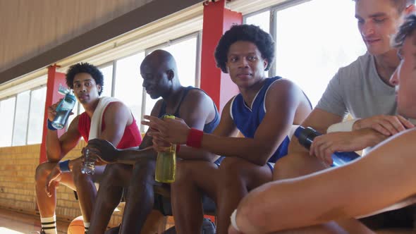 Diverse male basketball team and coach resting and drinking water after match