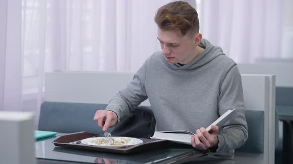 Hurrying Busy Caucasian Young Man Reading Book Eating Dessert in Canteen Indoors