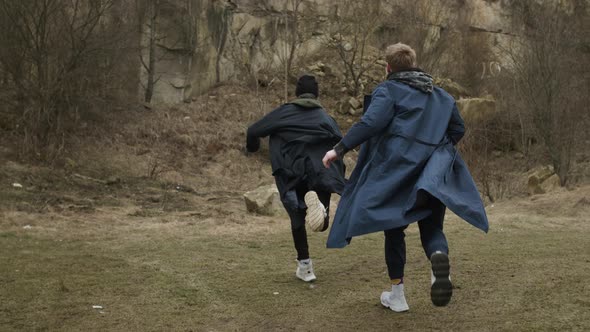 Two Young Guys Running Forward. Guys In Raincoats On Rock Background. View From The Back.