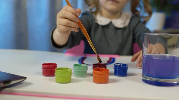 Child Schoolgirl Enthusiastically Drawing with Brushes Paints at Home Sitting at Table Using Laptop