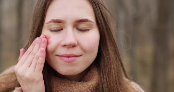 a fair-haired young girl applies liquid blush with her fingers on her face, smiling, rubs them again