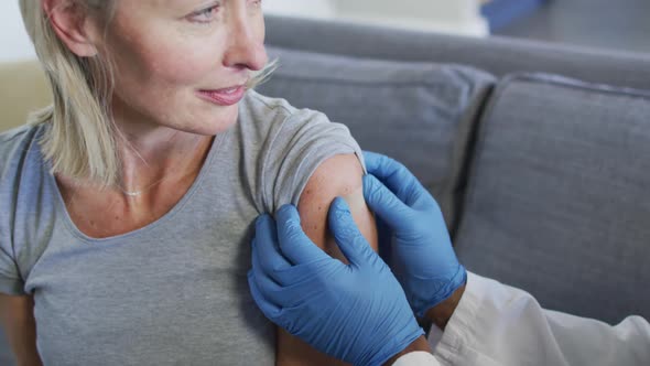 Happy senior diverse woman and doctor in living room sitting on sofa, vaccinating