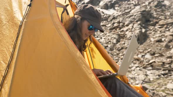 Lady Types on Laptop Sitting in Yellow Tent Against Hillside