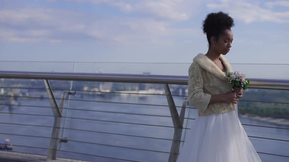 Frustrated Beautiful African American Woman in Wedding Dress Putting Bridal Bouquet on Bridge