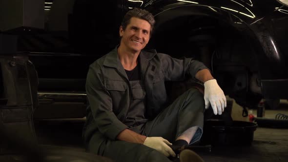 Waist up portrait view of the auto mechanic wearing gloves sitting near car outdoors