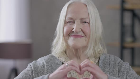 Old Greyhaired Caucasian Woman Showing Heart Shape with Hands Looking at Camera Smiling