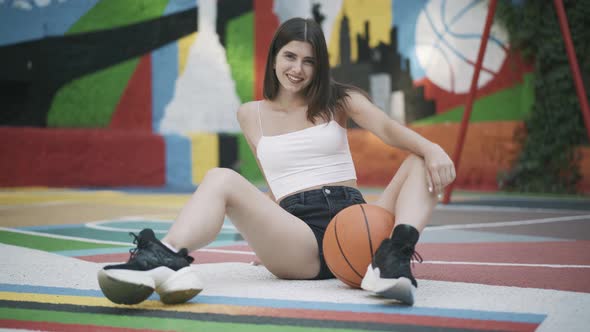 Smiling Confident Female Basketball Player Sitting on Outdoor Court with Orange Ball and Looking at