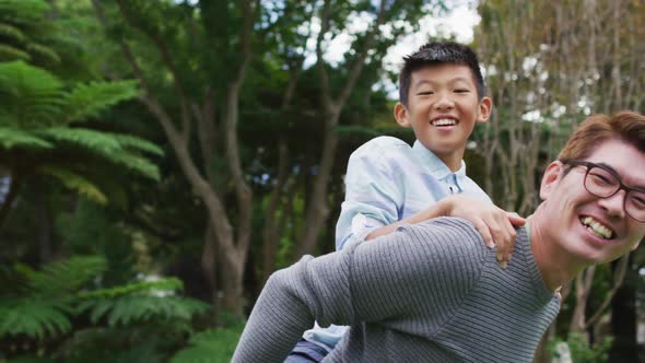 Smiling asian father piggy backing happy son having fun in garden together