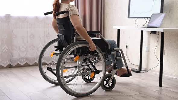 A Caucasian Woman in a Wheelchair Drives Up to Her Desk and Starts Typing with a Laptop From Home