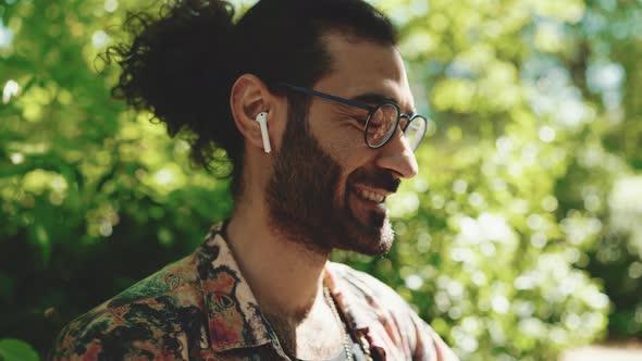 Positive curly-haired bearded man in eyeglasses listening music in headphones