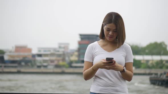 Young Asian woman using smartphone enjoying reading social media beside the fence at the river.