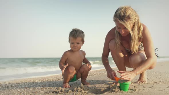 Mom with Glasses Plays with a Little Toddler Sitting on the Beach Near the Sea