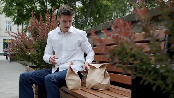 Man Sits on Street Bench with Smartphone Unwraps a Package with a Hamburger