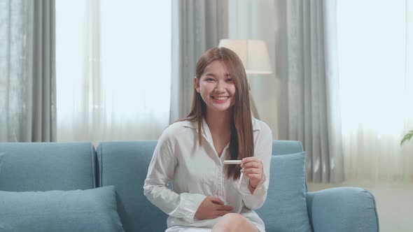 Happy Young Asian Woman Looking At Pregnancy Test While Touching Her Belly And Smiles To Camera