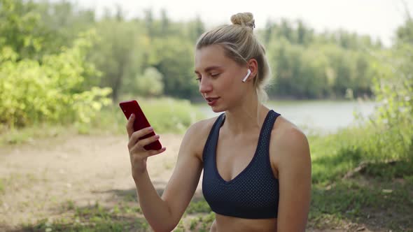 Woman in a Sports Top and Wireless Headphones Communicates on Phone Smiling in Nature. A Young Woman