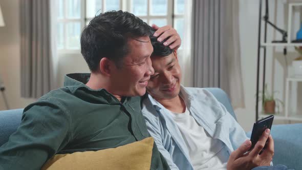 Asian Gentle Gay Couple Using Mobile Phone While Sitting On A Couch, Watching Funny Videos