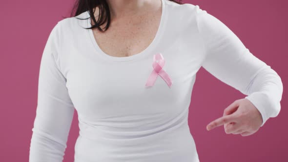 Mid section of a woman pointing to the pink ribbon on her chest against pink background