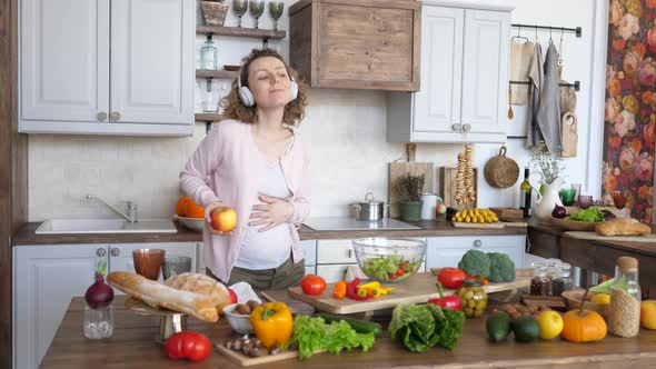 Pregnant Woman Wearing Headphones And Dancing With Apple On Kitchen
