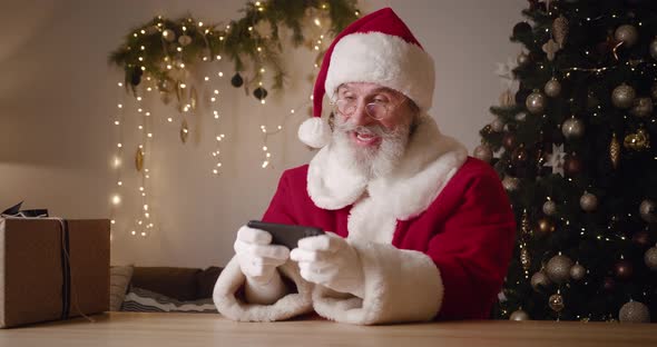 Joyful Elderly Santa Claus in a Carnival Red and White Suit with Hat Sits Against the Background of