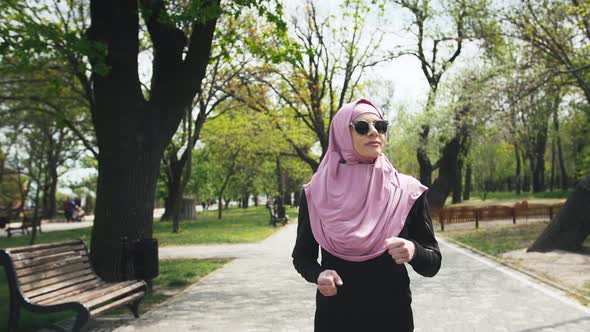 Portrait of Young Modern Muslim Woman in Sunglasses and in Hijab Jogging in Green Park