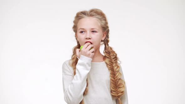 Young Adorable Caucasian Girl with Long Pretty Blonde Hair Eating Apple and Smiling on White