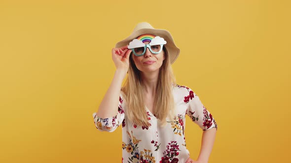 Comical Fairhaired Woman in a Floral Blouse and a Beige Hat Taking Off Extravagant Rainbow Glasses