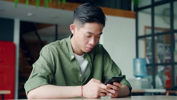 Young Happy Focused Asian Student Sitting at Office Using Mobile Cell Phone