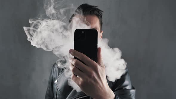 Smoker Man in Leather Jacket Hide His Face with Smartphone and Blowing Smoke Over Dark Grey