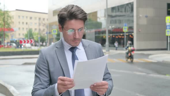 Middle Aged Businessman Reading Documents while Walking on Street