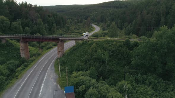 An Abandoned Rusty Railroad Bridge Above the Curving Road with a Driving Truck