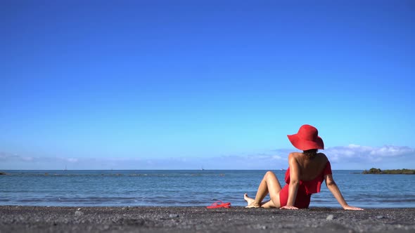 Woman in a red swimsuit and hat sits on a black sand beach. Canary Coast