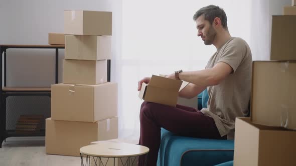 A Young Man Opens a Cardboard Box and Takes Out Things Sitting on the Sofa in a New Apartment After
