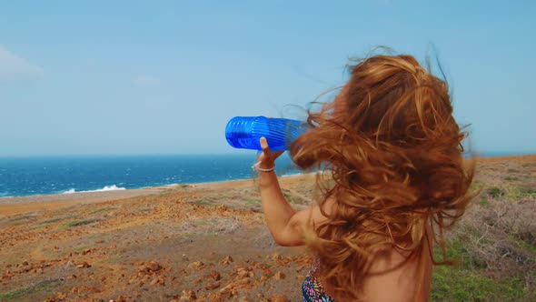 Beautiful woman drinking from blue water bottle by ocean with wind blowing hair