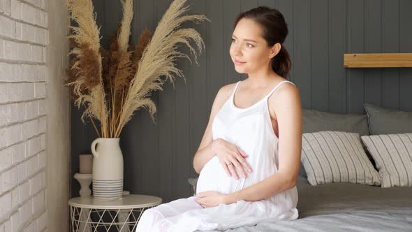 Pregnant Lady in White Dress Strokes Tummy Sitting on Bed